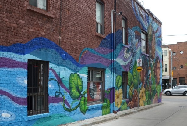 looking down a short narrow alley with a large colourful mural on the left side, blue background with leaves and vines in the foreground