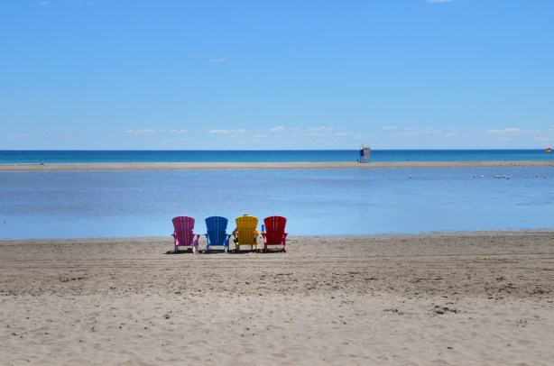 4 muskoka chairs lined up on the beach, pink, blue, yellow, and red, lots of water, a person is sitting in one of them but photo is of back of chairs. 