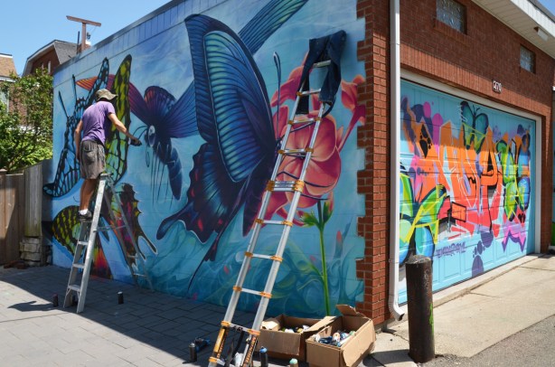 Nick Sweetman on a ladder adding the finishing touches, with spray paint, to a large mural of butterflies on the side of a garage in a lane. Another ladder leans against the same wall, boxes and can of spray paint on the ground. On the same garage, there is a colourful mural on the garage door 