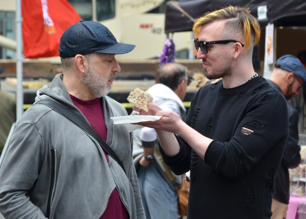 a man with glasses and hair that is shaved on one side of his head is offering another man a rice krispie square, he is holding it to the man's mouth