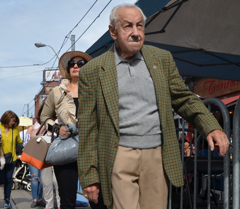 an older man walking on the street with a woman behind him in a large straw hat and dark sunglasses and carrying a large brown and orange purse as well as a grey plastic bag 