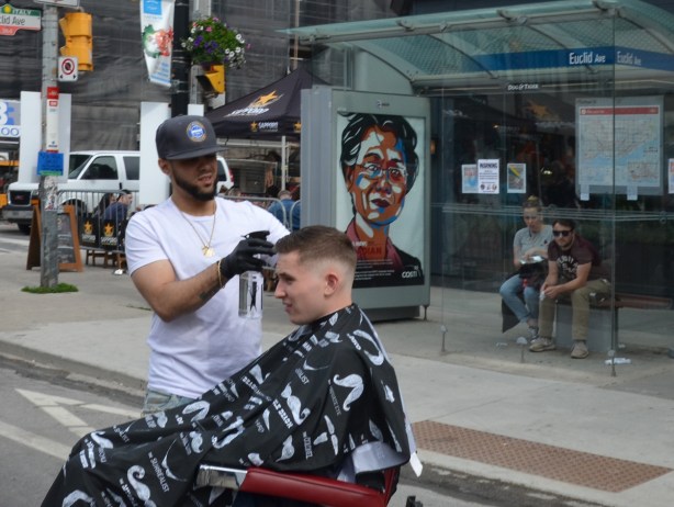on the street, a man is giving another man a haircut, two people are sitting on a bench in a bus shelter behind, watching 