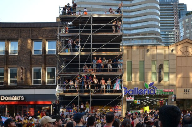 people sitting onscaffolding in the front of a three stprey building on Yonge street, watching the pride parade 