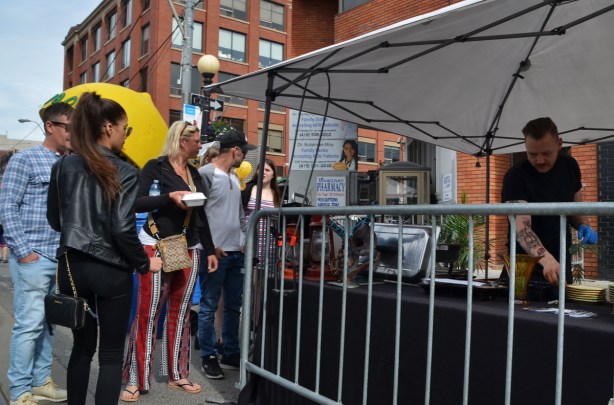 people in a line up to get food at an outdoor vendor, white tent covering, 