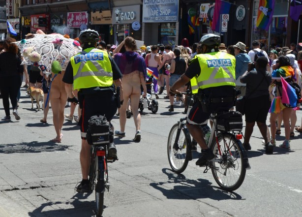 two police on bikes with yellow vests ride in parade behind two people with bare bottoms