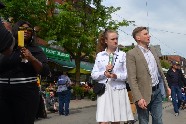 a woman with a yellow popsicle, a woman in a white skirt and top and a man in a beige suit jacket, all walking on the street 