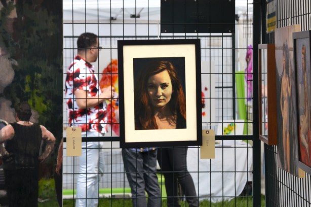 a framed portrait of a woman on a metal grid, people standing behind it including a man with a white shirt with bright red and black blotches