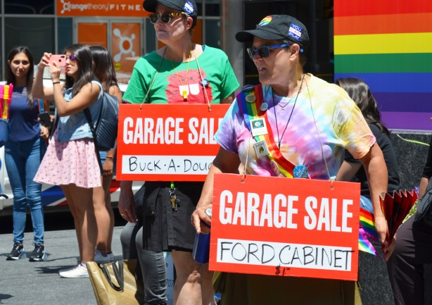 two people at dyke march with anti Doug Ford protest signs, garage sale signs, Ford cabinet for sale for 10 dollars