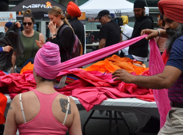 the back of a woman with white bra straps showing and a large tattoo on ther shoulder. A man pulls a length of pink fabric as he wraps her head in a pink turban