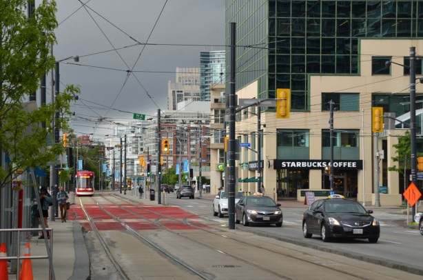 looking west on Queens Quay at Spadina, streetcar tracks, street, trafiic, pedestrian on sidewalk, TTC street car approaching, Starbucks on the corner, low rise buildings in the background 