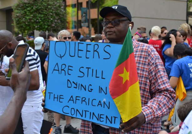 a black man holds a sign that says Queers are still dying in African continent, he is holding a flag from Cameroon 