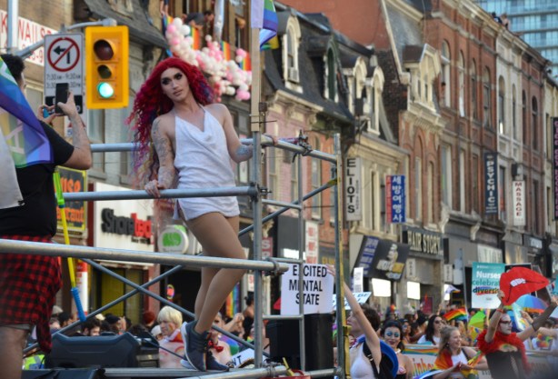 a person in a long red wig and a short white dress is posing for a male photographer on a float in the pride parade, people walking behind the float 