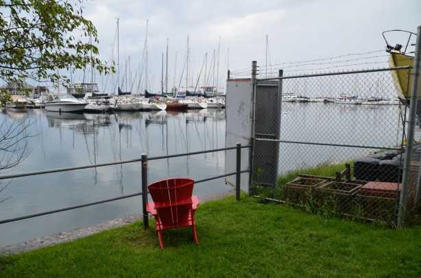 a lone red Muskoka chair sits on an angle in a small grassy patch between a fence and a path, looks out over a yacht club 