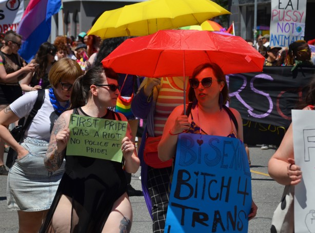 women walking in dyke march,under red umbrella with a sign that says bisexual bitch, woman beside her has a small sign that says first pride was a riot, no police