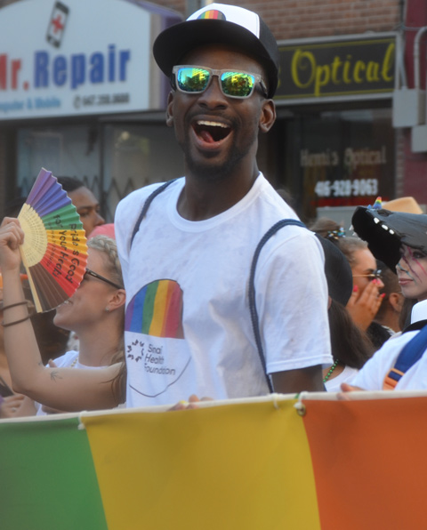 a young black man with black baseball cap and green sunglasses has a big smile as he helps carry a banner in the pride parade 