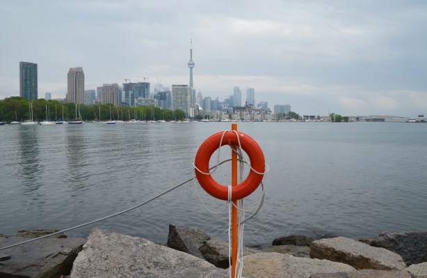 In the background is the Toronto skyline from Trillium Park, from the green trees of Coronation Park on the left and past the CN Tower and tall city center buildings, to the National Yacht Club and then Billy Bishop airport on the far left. In the foreground is an orange lifesaving ring and the rocks of the shoreline of the park