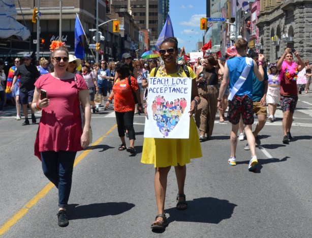 women walking in dyke march, one woman in a bright yellow dress carrying a sign that says teach love and acceptance, with a heart made of people