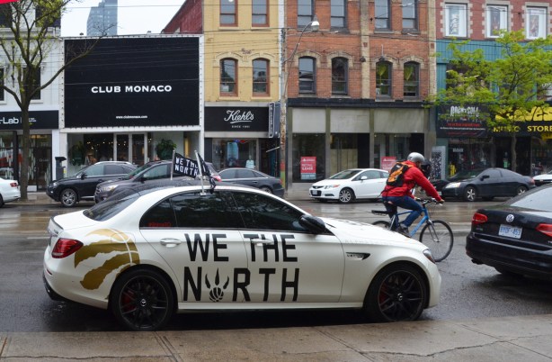 a white car is parked by the side of the road, it is decorated withlarge black letters that say We the North, it is also flying a black and white flag witgh the same words on it 