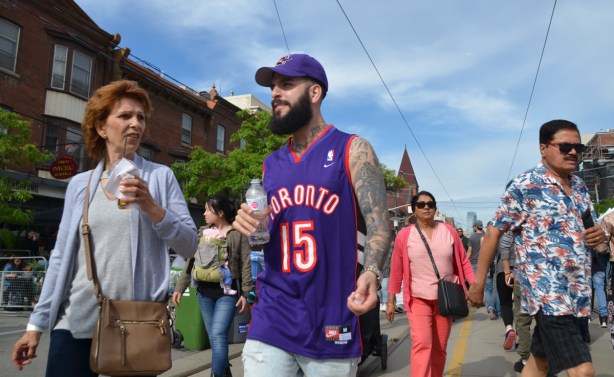 people walking down the street towards the camera, a man in a purple Raptors shirt with marching baseball cap, a woman in a light blue blouse with a drink in her hand, a woman in a pink pant suit 