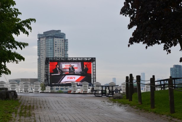 a large screen plays a TSN show, outdoors, Ontario Place, white chairs but no one is there 