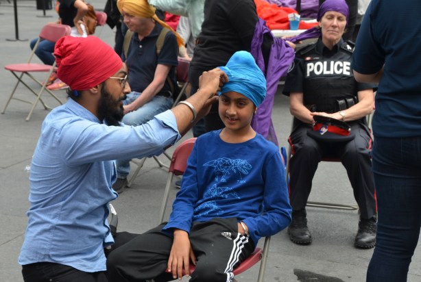 At Turbanup event at Yonge Dundas Square, a man in beard and red turban wraps the head of young person in a blue turban, in the chair behind is a female police officer having her head wrapped in a purple turban