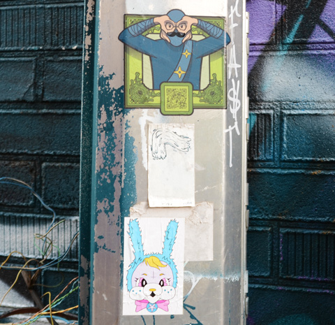 three stickers and paste ups on a metal post, urban ninja warrior on the top, with a black mustache, and a pink and blue bunny face on the bottom 