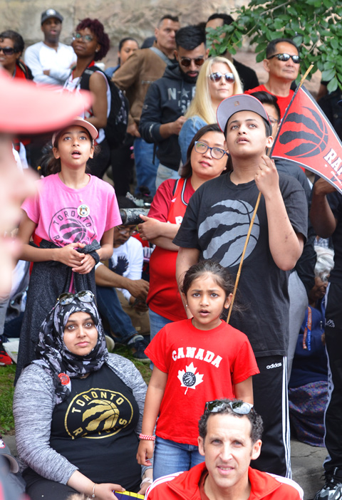 Raptors fans, parade day, sitting on street and sidewalk, watching a video screen showing the parade as it makes its way towards Nathan Philips Square 