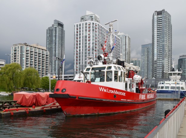 The WIlliam Lyon McKenzie, a bright red fire boat is docked at Toronto waterfront, city skyline behind the boat with tall condos, also dark storm clouds 