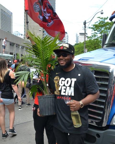 Raptors fans, parade day, a man with a bottle of white wine, and a large potted palm plant, standing in front of a truck, a large red RAptors fly is being held behind him 