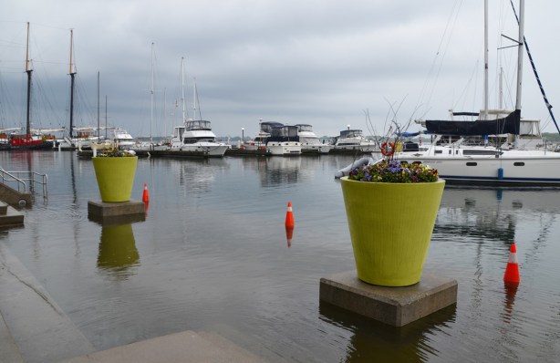 two large light green flower pots sit on concrete pedestals, in the water, orange cones in the water, shoreline is flooded, some sailboats docked in the background 