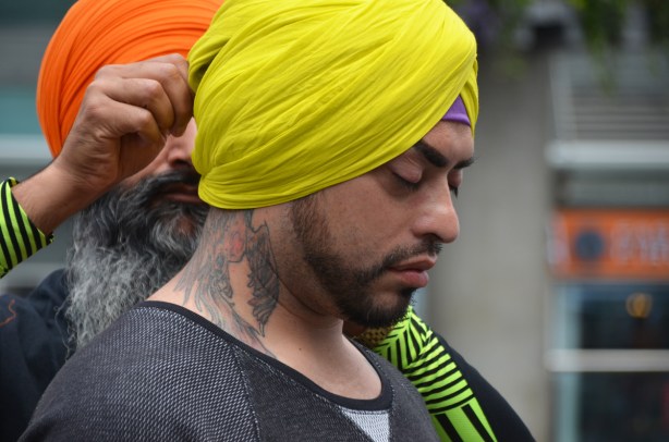 an older man with a beard and an orange turban, is tying a yellow turban onto the head of another man who has a large tattoo on his neck