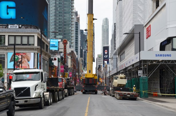 a large crane is on a truck in the middle of Yonge street, downtown, with tall buildings on both sides including the Eaton Centre under renovation on the right