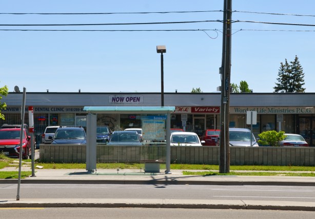 looking across the street to a bus shelter that is in front of a strip mall with cars parked in front of the stores 