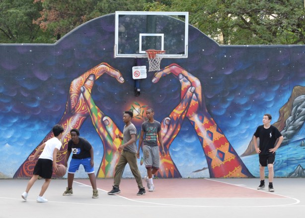 a group of boys playing basketball on a court that has a mural of two hands forming a heart shape with their hands, the heart is under the basket, mural is on wall
