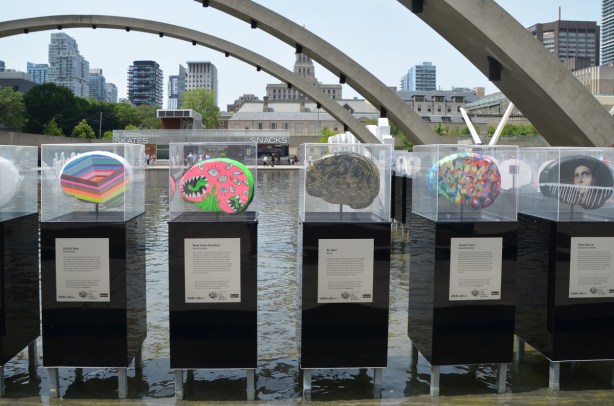 five brain shaped sculptures in plexiglass display boxes standing in the pool at Nathan Phillips Square