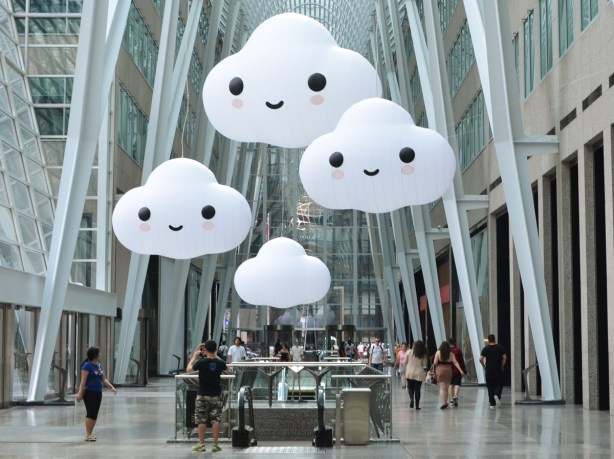 art installation in Brookfield Place of 4 large white clouds, three of which have happy faces on them, suspended from the ceiling over the escalator from the lower level,