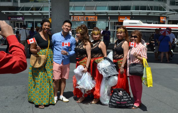 a group having their picture taken at Yonge Dundas Square, one Asian man and four women. Three women are in costume as they are about to perform on the stage in that square, Canada Day celebrations