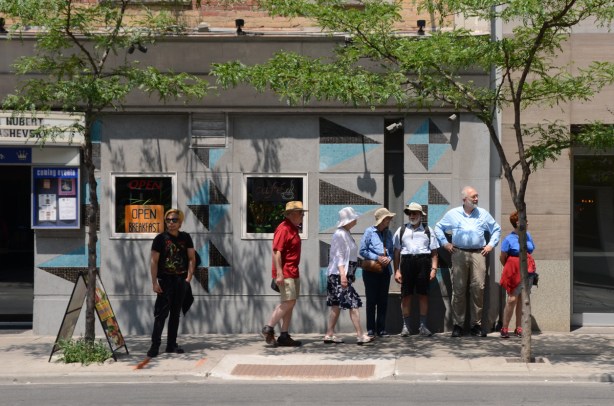 a group of older men and women in sun hats stands in a bit of shade cast by a little tree outside the Rex on Queen Street West 