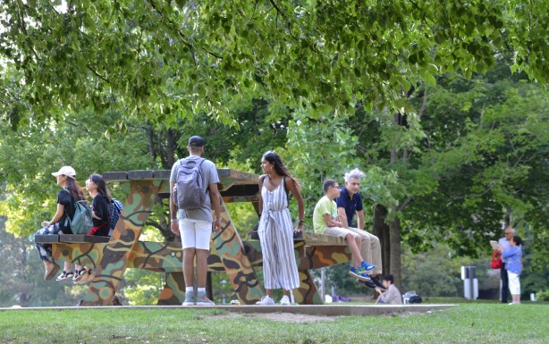 a group of people sit on an oversized picnic table painted in camo colours in a park 