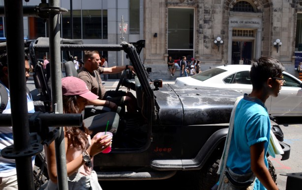 a man in an open jeep is driving down Yonge Street, people walking are passing him as he is stopped for a red light 
