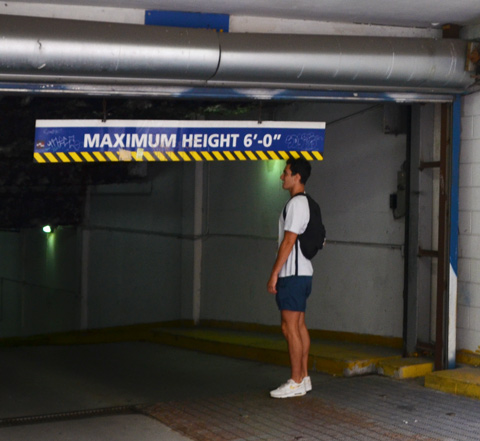 a young man stands by a maximum height 6 feet sign at the entrance of a parking garage, to see if he is 6 feet tall 