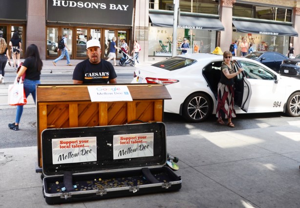 Mellow Dee, a male musician, plays the piano outside Eaton Centre on Queen street as a woman gets out of a white taxi behind him 
