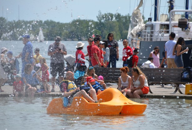 people in a yellow plastic paddle boat on a man made pond near the waterfront, a fountain is spraying them, they are paddling past a group of people on sitting on the edge of the pond 