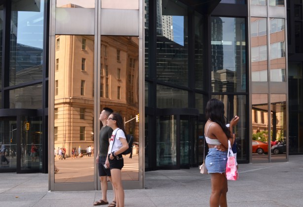 three people in front of the R B C building on Front St.., with its gold coloured reflective windows, An Asian couple stopped to look up and a black woman taking a picture, reflections of other buildings nearby. All people are wearing shorts