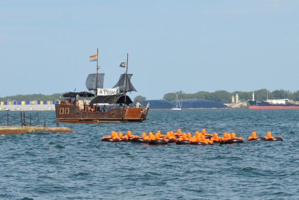 a pretend pirate ship, as a harbour cruise boat passes by the public art installation, SOS, or Safety Orange Swimmers