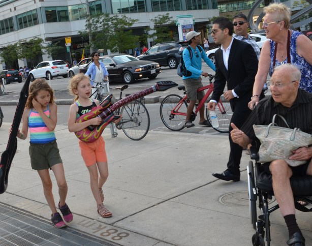 two young girls pretending to make music with large plastic inflatable guitars while a woman pushing a man in a wheelchair look on.