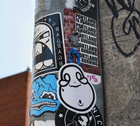 slaps stickers on a pole, top one is Portland is burning with man pasting sign to wall, another has a man with a fat chin and mouth open in O shape. 