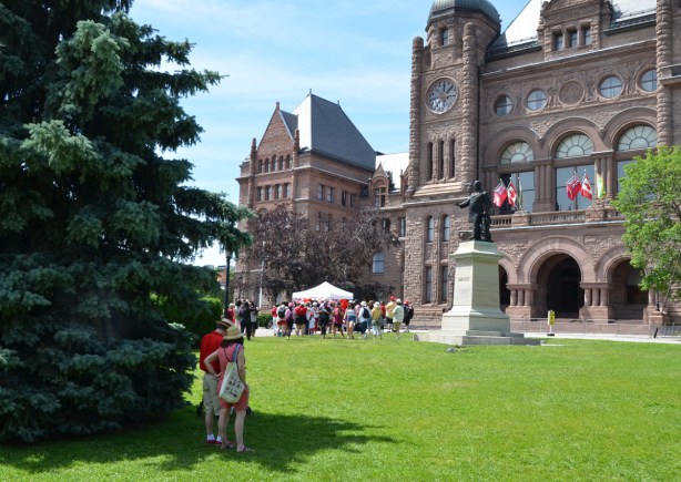 front of Queens Park buildings, parliament buildings, on the grass a couple stand by a tree, looking at group under a tent, Canada Day celebrations 