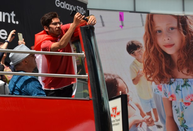 a red head girl in a large advert on a building beside a man in red adjust a microphone on the top of a red double decker tourist hop on hop off bus 