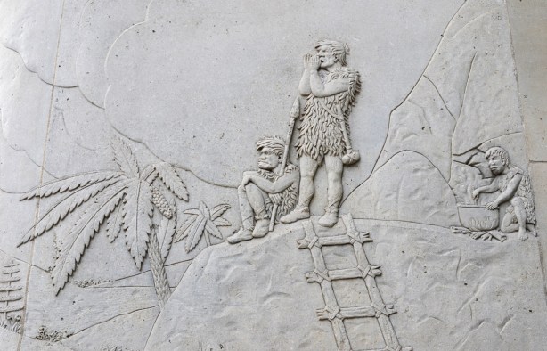 relief sculpture in concrete on exterior of wall, cavemen scene, with palm trees, three people dressed in animal skins. One is cooking - stirring with a stick in a large pot over a fire, one is standing and shouting with hands cupped around his mouth. A ladder made of wood pieces lashed together leans against a rock 